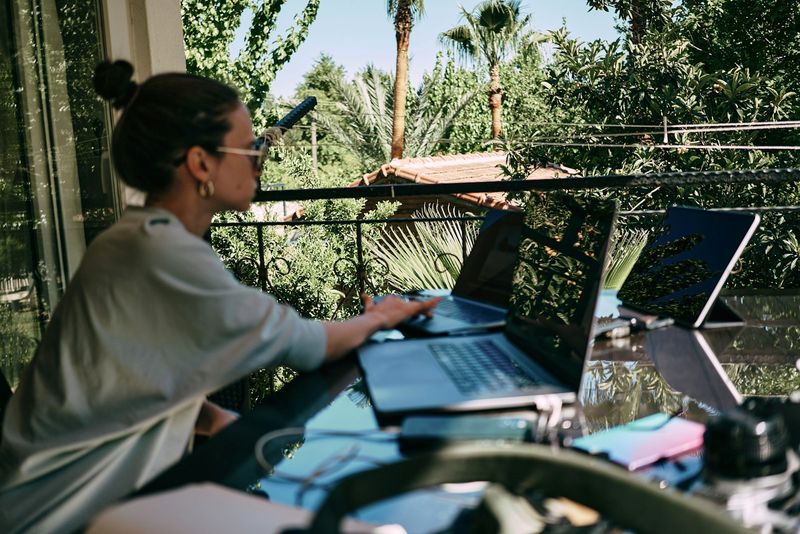 A freelancer working on a laptop at a sunny terrace table among palm trees